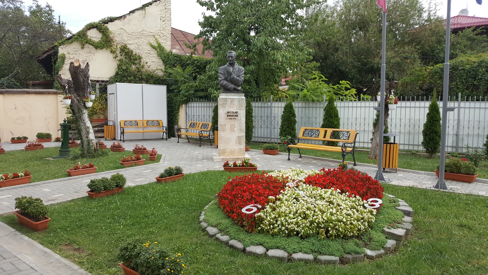 Grădina muzeului, cu bustul lui Nicolae Simache, bănci și un ceas din flori roșii și albe, cu cifre pe margine