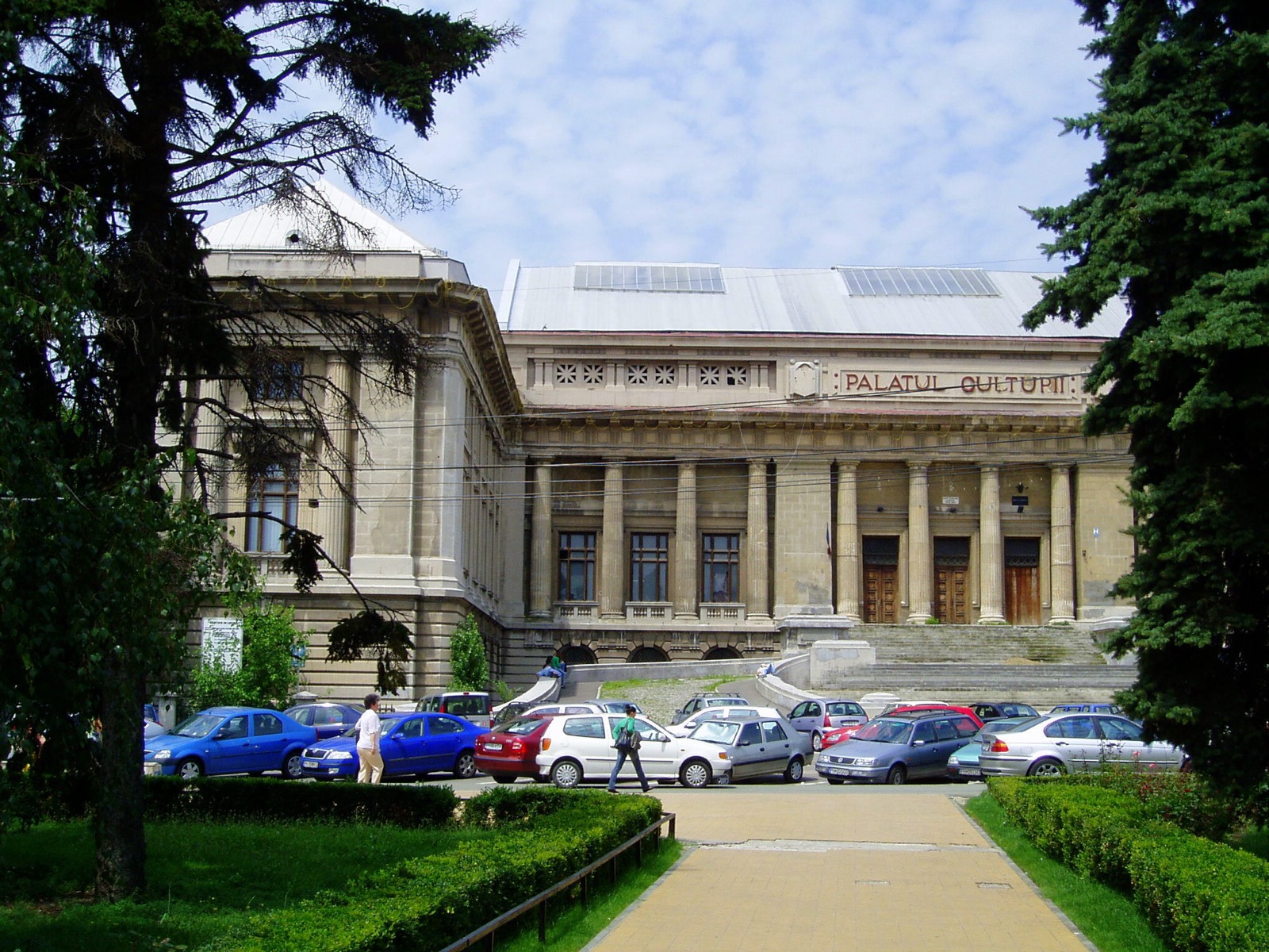 Palatul Culturii văzut din parcul din față, între brazi, cu mașini parcate în fața colonadei